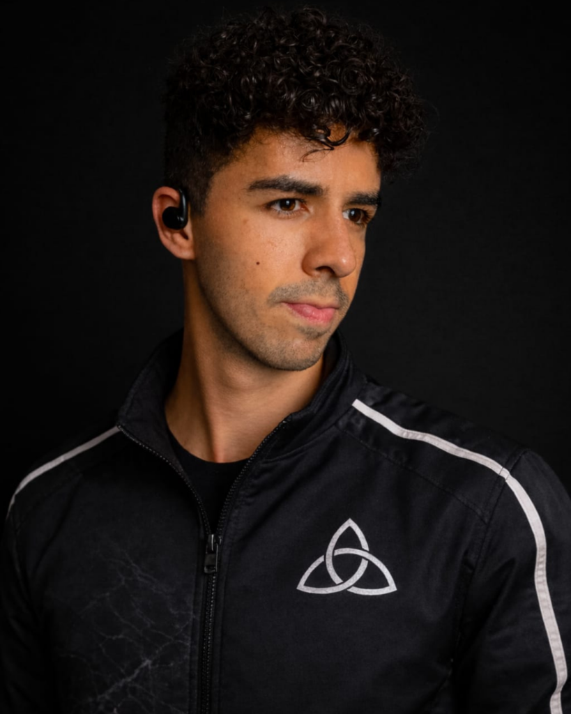 Simon Aleckovith, o criador do app Multiverso posando para foto oficial e vestindo o uniforme de sua empresa, com a logomarca que representa o símbolo milenar na triquetra.